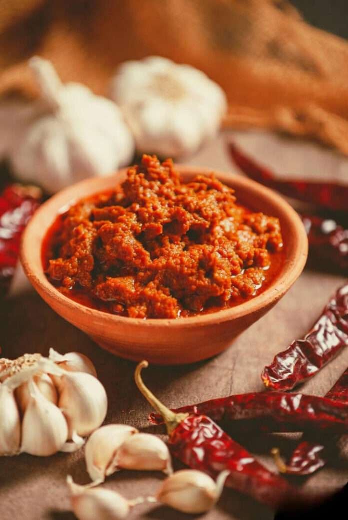 a clay bowl filled with bright red Rajasthani lahsun ki chutney, with a few whole garlic pods and dried red chilies scattered like beautiful chaos on a rustic table. a clay bowl filled with bright red Rajasthani lahsun ki chutney, with a few whole garlic pods and dried red chilies scattered like beautiful chaos on a rustic table.