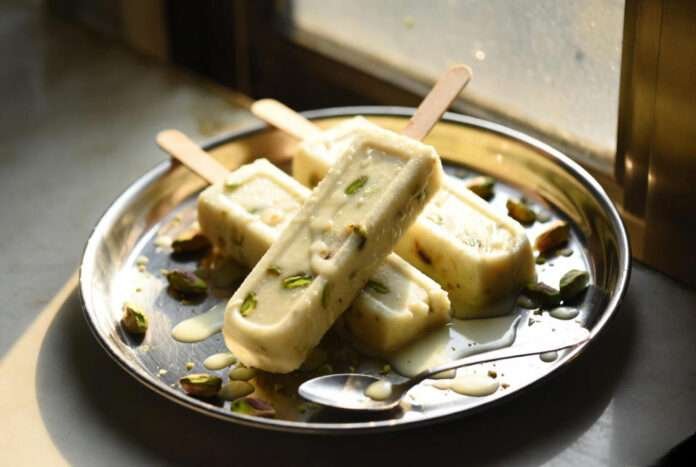 three kulfi sticks placed on a steel plate with melting drips pooling around them.